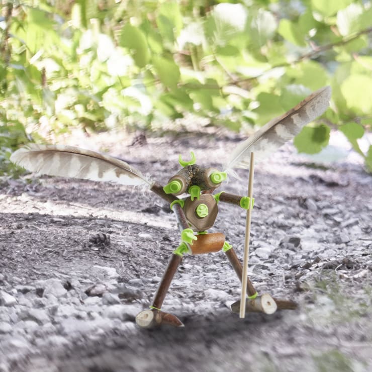 Eine kleine Figur aus Zweigen, Blättern, Samenkapseln und Federn steht auf einem unbefestigten Weg und hält einen Stock in einer Hand, während das grüne Laub im Hintergrund verschwimmt.