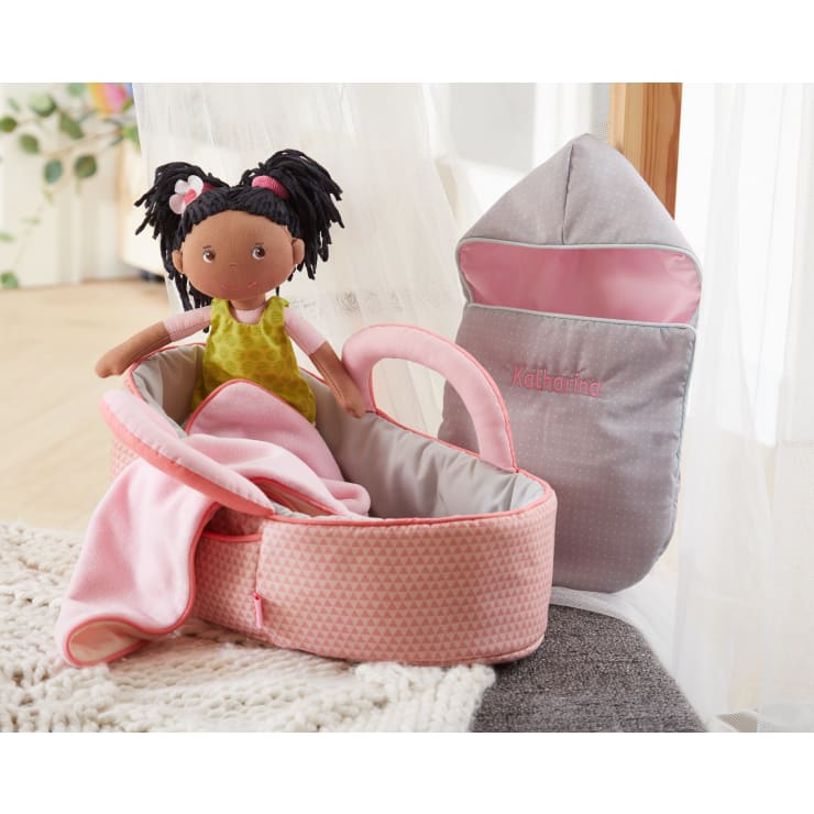 Eine weiche Puppe mit dunklen Zöpfen und einem grünen Kleid sitzt in einer rosa gemusterten Babytrage mit einer rosa Decke. Daneben liegt ein grau-rosa Babyschlafsack mit der Aufschrift Katharina.