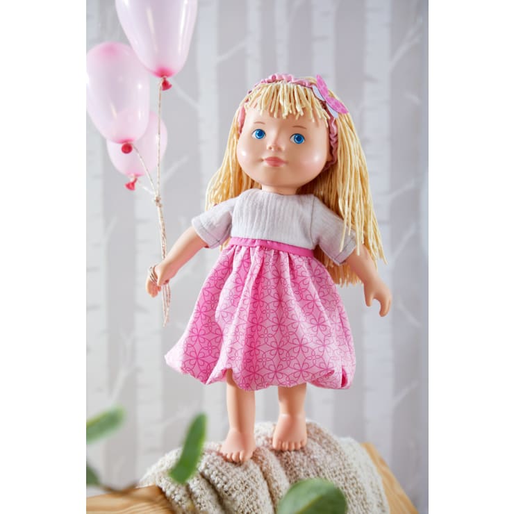 Eine blonde Puppe mit blauen Augen, die ein rosafarbenes Stirnband und ein rosafarbenes und weißes Kleid trägt, steht auf einer Decke und hält rosafarbene Luftballons in der Hand. Der Hintergrund besteht aus einem hellen Birkenmuster.
