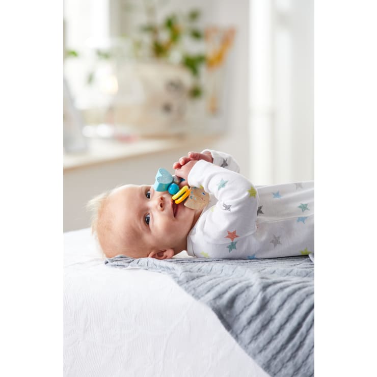 Ein Baby liegt auf dem Rücken auf einem Bett, hält ein buntes Beißspielzeug in der Hand und kaut darauf herum. Das Baby trägt einen weißen Strampler mit einem Sternenmuster. Der Hintergrund ist mit unscharfen Haushaltsgegenständen sanft beleuchtet.