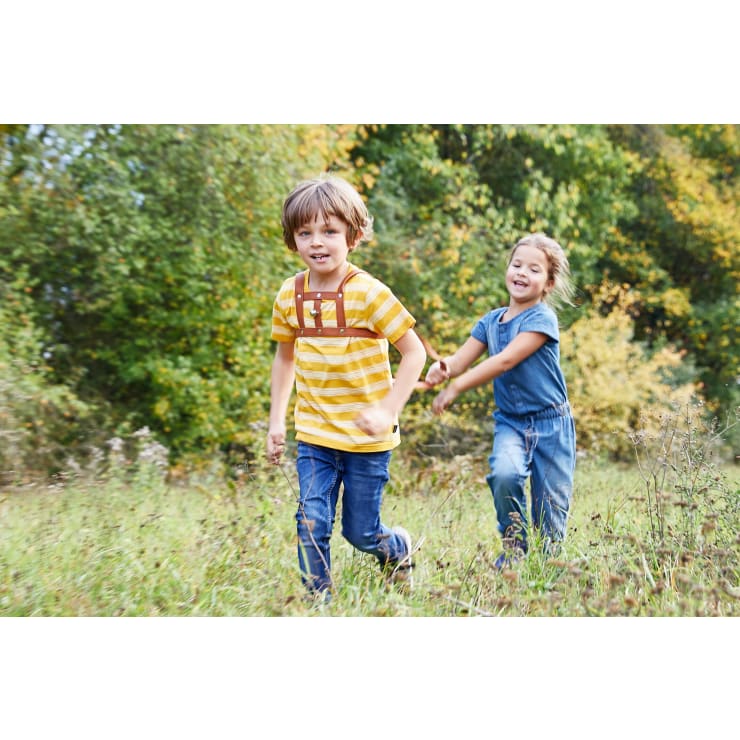 Ein Junge in einem gelben gestreiften Hemd rennt vor ihm her, während ein Mädchen in einem blauen Outfit ihn durch ein grasbewachsenes Feld mit grünen Bäumen im Hintergrund verfolgt. Beide Kinder scheinen im Freien zu spielen.