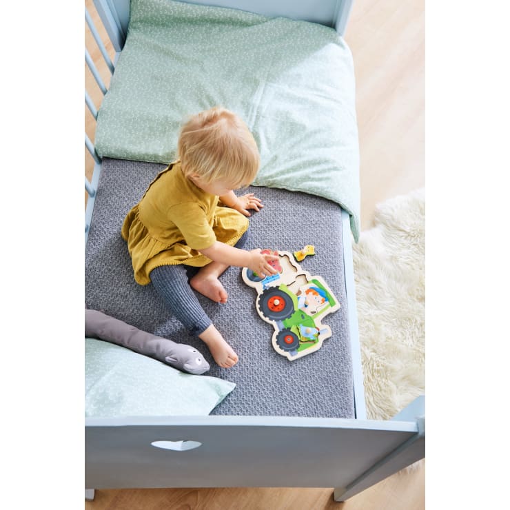 Ein kleines Kind mit blondem Haar sitzt auf einem Bett mit grünem Bettzeug und setzt ein Holzpuzzle in Form eines Traktors zusammen. Das Kind trägt ein gelbes Kleid und graue Leggings. Das Bett steht direkt neben einem Plüschteppich.
