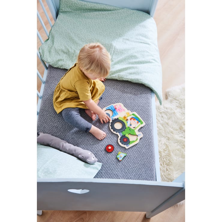 Ein kleines Kind mit blondem Haar sitzt in einem Kinderbett auf einer grauen Matte, trägt ein gelbes Kleid und gestreifte Leggings und spielt mit einem bunten Holzpuzzle. Ein grünes Kissen, eine Decke und ein Plüschtier liegen in der Nähe.