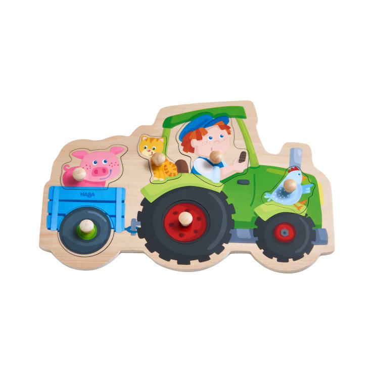 Ein Holzpuzzle mit einem grünen Traktor, der von einem Kind gefahren wird, mit einer Katze und einem Schwein, die in einem blauen Anhänger sitzen, und einem Vogel, der auf dem Traktor thront. Jedes Teil hat einen Holzknauf für eine einfache Handhabung.