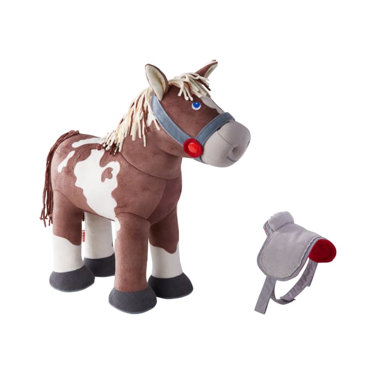 Ein braun-weißes Plüschpferd mit grauem Zaumzeug und blauen Augen steht neben einem abnehmbaren grauen Sattel mit rotem Futter, beides auf weißem Hintergrund.