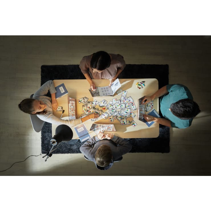 Vier Personen sitzen um einen rechteckigen Tisch, der mit Spielkarten und Heften bedeckt ist, und beschäftigen sich unter einer Lampe mit Karten, die von oben auf einen Holzboden mit einem dunklen Teppich darunter gerichtet ist.