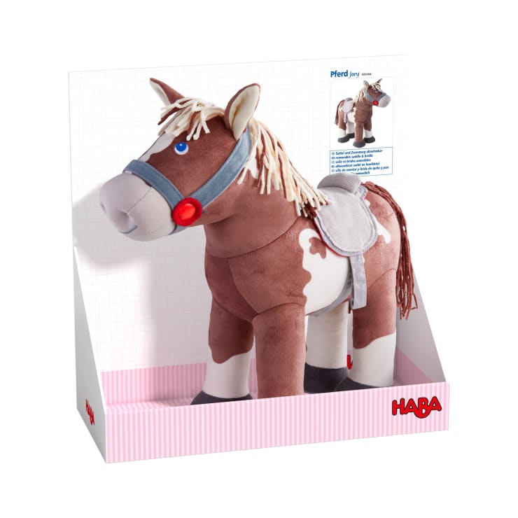 Ein Plüschpferd mit braun-weißem Körper, beiger Mähne und Schweif, grauem Zaumzeug und Sattel, in einer weiß-rosa gestreiften Schachtel mit HABA-Logo in der rechten unteren Ecke.