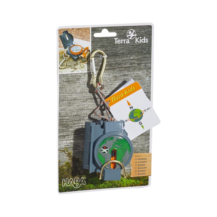 Ein verpackter Terra Kids Kompass von HABA mit grauem und grünem Design, Metallkarabiner und beiliegender Anleitungskarte. Die Verpackung zeigt den Produktnamen und ein Foto des Kompasses im Gebrauch auf einem Holzhintergrund.