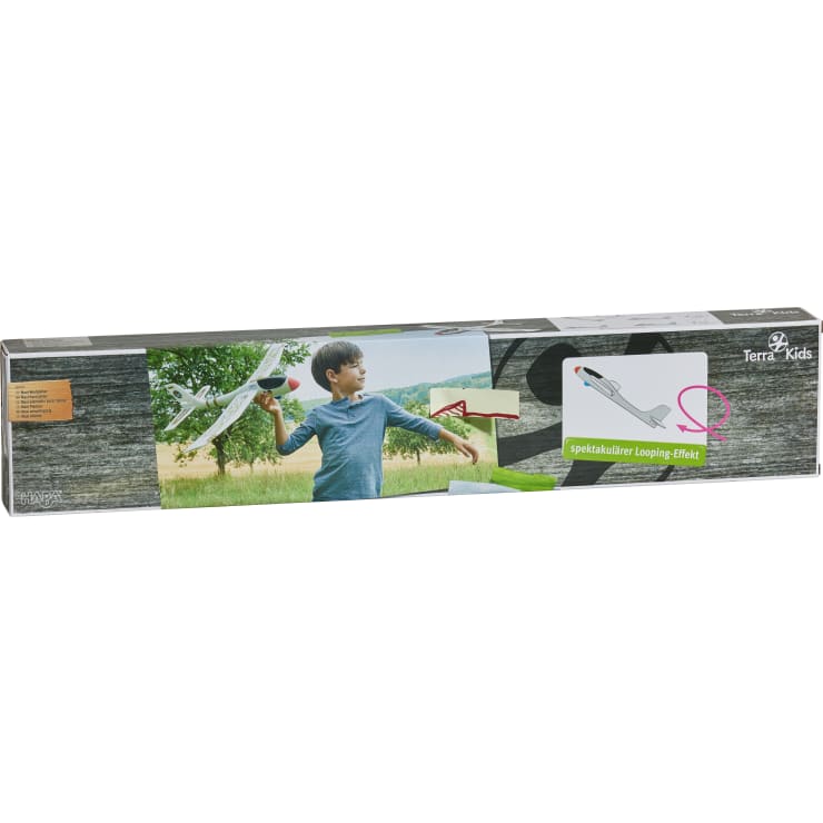 Ein kleiner Junge hält im Freien ein weißes Spielzeugflugzeug aus Schaumstoff mit schwarzen und roten Akzenten. Das Bild befindet sich auf einer rechteckigen Schachtel mit der Aufschrift Terra Kids. Auf der Verpackung sind auch Illustrationen der Bewegung des Flugzeugs und ein Logo zu sehen.