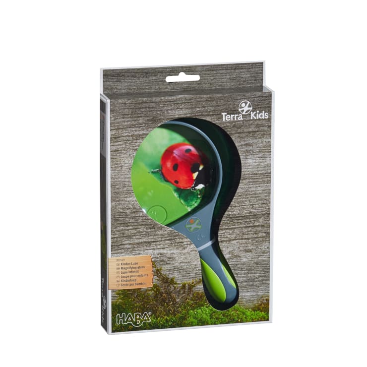 Eine Handlupe in einer schwarz-grünen Verpackung mit der Aufschrift Terra Kids von HABA, auf der ein Marienkäfer in Großaufnahme auf einem Blatt abgebildet ist. Die Verpackung ist vor einem hölzernen Hintergrund angebracht.