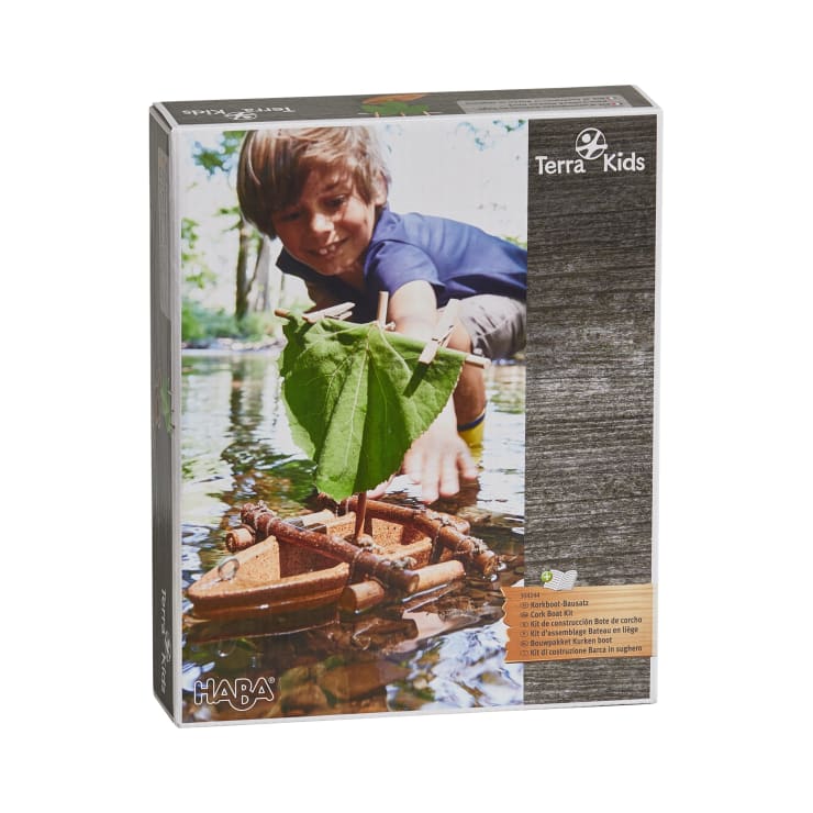 Auf einer Schachtel des HABA Terra Kids Kreativbaukastens ist ein Kind abgebildet, das ein kleines Holzfloß mit einem grünen Blattsegel in einem seichten, begrünten Bach zu Wasser lässt.
