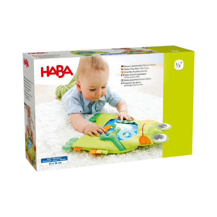 Ein Baby liegt auf einem weißen Teppich und spielt mit einer grünen Froschmatte. Auf der Produktverpackung ist die Marke HABA abgebildet und es wird angegeben, dass es für Kinder ab 6 Monaten geeignet ist. Auf dem Karton sind Produktdetails in mehreren Sprachen abgebildet.