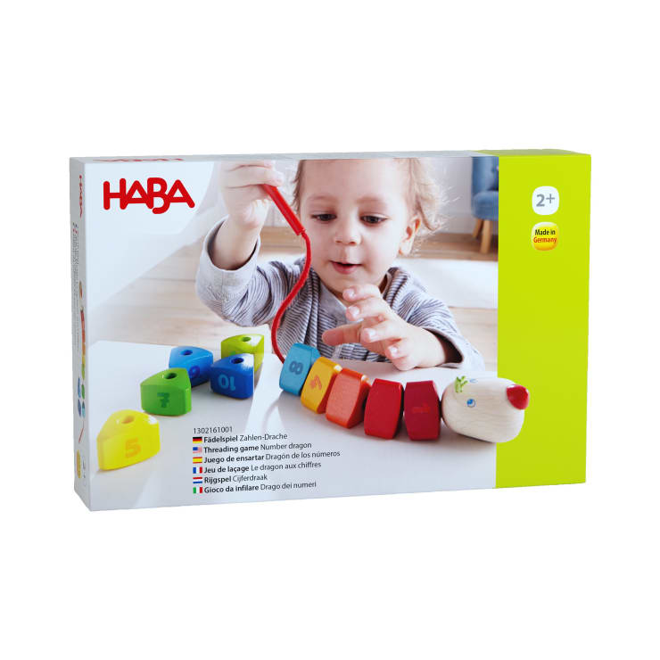Ein kleines Kind fädelt bunte, nummerierte Holzklötze auf eine rote Schnur neben einem hölzernen Drachenkopf. Die Szene ist auf der Verpackung eines HABA-Spielzeugs abgebildet, das für Kinder ab 2 Jahren empfohlen wird.
