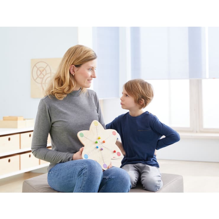 Eine Frau in einem grauen Rollkragenpullover hält eine hölzerne Lehrtafel mit farbigen Wäscheklammern, während sie neben einem Jungen in einem blauen langärmeligen Hemd sitzt. Sie sitzen in einem Raum und schauen sich an.