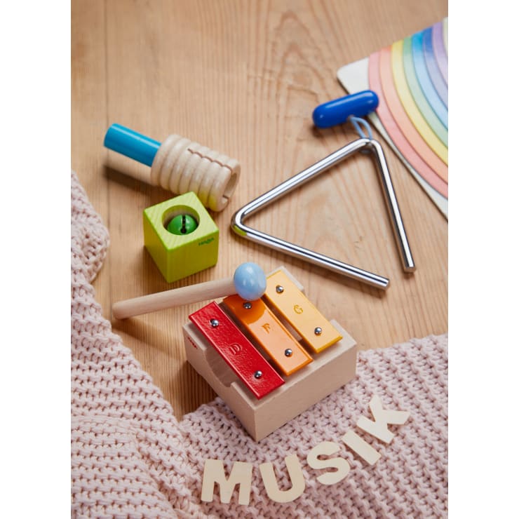 Musikinstrumente für Kinder, darunter ein Xylophon, eine Triangel, ein grüner Glockenschüttler und ein Rhythmusstab mit blauem Griff, sind auf einer hölzernen Fläche mit einem gestrickten Tuch und Holzbuchstaben angeordnet, auf denen "MUSIK" steht.