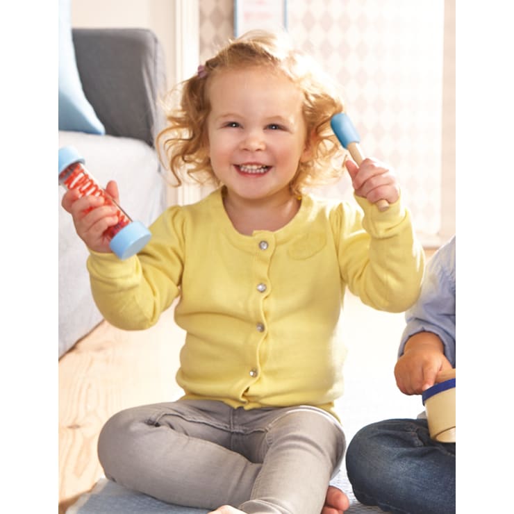 Ein kleines Kind mit lockigem Haar, das eine gelbe Strickjacke und eine graue Hose trägt, sitzt im Haus auf dem Boden und lächelt, während es zwei blaue und orangefarbene Musikshaker in jeder Hand hält.