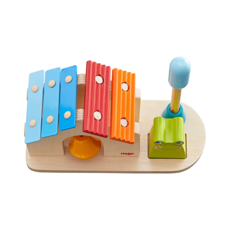 Ein Holzspielzeug mit einem Xylophon mit blauen und roten Tasten, einem grünen froschförmigen Block, einer gelben Glocke und einem blauen Schlägel auf einem hellen Holzsockel.