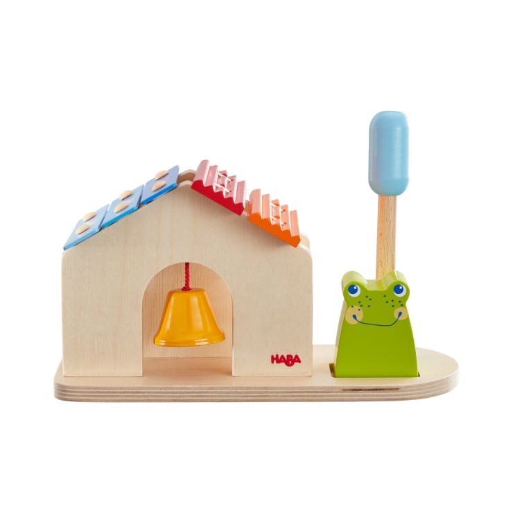 Ein Holzspielzeug mit einem kleinen Haus mit einer gelben Glocke im Inneren, einem Xylophondach mit bunten Stäben und einem lächelnden grünen Frosch auf der rechten Seite, der einen blauen Schlägel hält. Die Marke HABA ist auf dem Haus aufgedruckt.