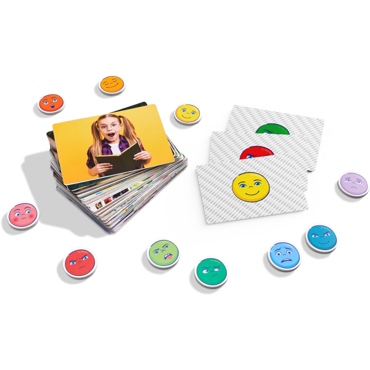 Ein Kartenstapel mit einer Karte, die ein überraschtes Mädchen mit einem Buch in der Hand zeigt, drei verdeckte Karten mit Smiley-Gesichtern und mehrere runde Spielsteine mit verschiedenen Gesichtsausdrücken umgeben die Karten.