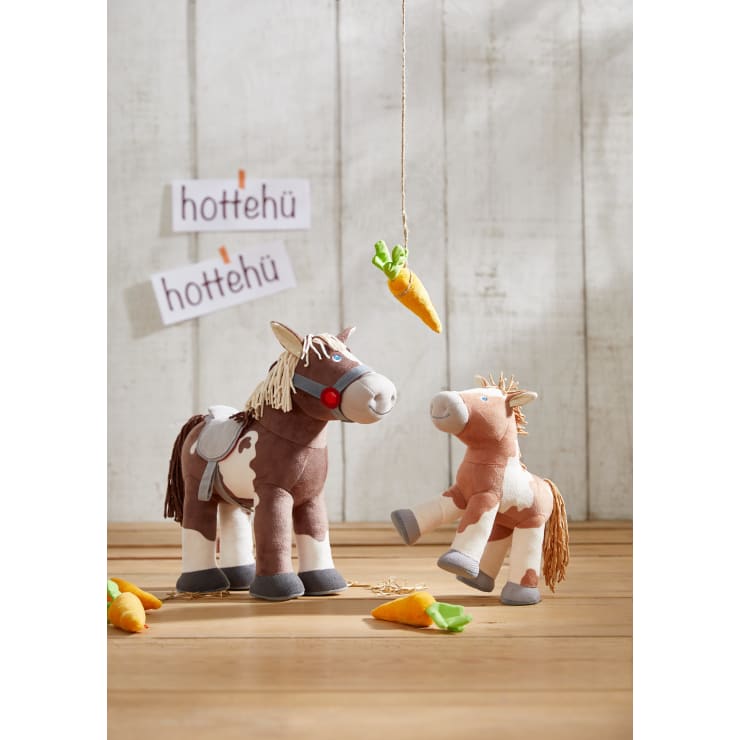 Zwei Plüschpferde stehen auf einem Holzboden und sind von Stoffmöhren umgeben. Darüber hängt eine Karotte an einer Schnur. An einer Holzwand im Hintergrund sind Schilder mit der Aufschrift "hottehü" angebracht.
