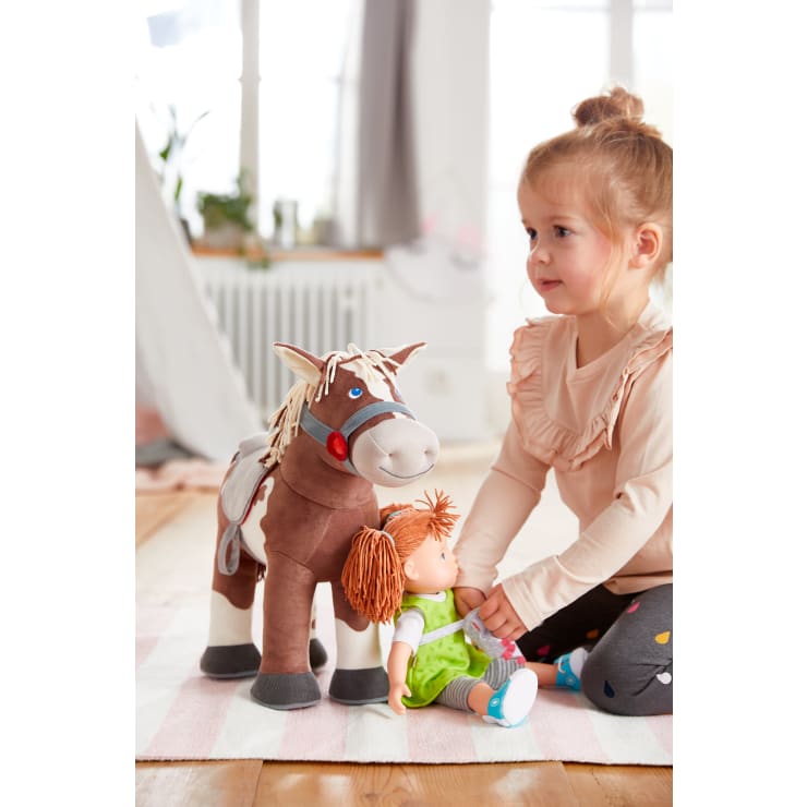 Ein junges Mädchen sitzt auf einem Teppich in einem Haus und hält eine grün gekleidete Puppe in der Hand. Neben ihr steht ein großes Plüschpferd mit einem Sattel und Zügeln. Der Raum ist hell mit natürlichem Licht und im Hintergrund stehen Pflanzen.