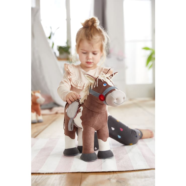 Ein kleines Kind sitzt auf einem gestreiften Teppich im Haus und spielt mit einem großen braunen und weißen Stoffpferd. Das Kind trägt ein helles Rüschenhemd und eine dunkle Hose mit bunten Punkten. Im Hintergrund sind Pflanzen und sanftes Licht zu sehen.