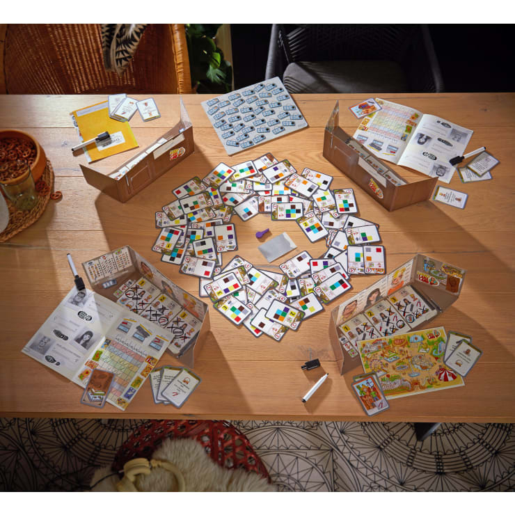 Auf einem Holztisch sind Spielbretter, Karten, Spielsteine und Trockenlöschmarker ausgebreitet. In der Mitte befinden sich vier Spielerstationen, eine Anleitung und verschiedene bunte Spielfiguren.