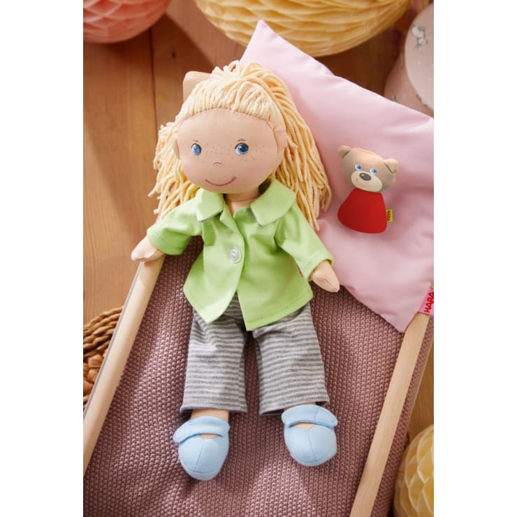 Eine Stoffpuppe mit blondem Garnhaar, grünem Hemd, gestreifter Hose und blauen Schuhen liegt auf einem rosa Kissen mit kleinem Bärenmotiv und einer lila Decke auf einer Holzfläche.