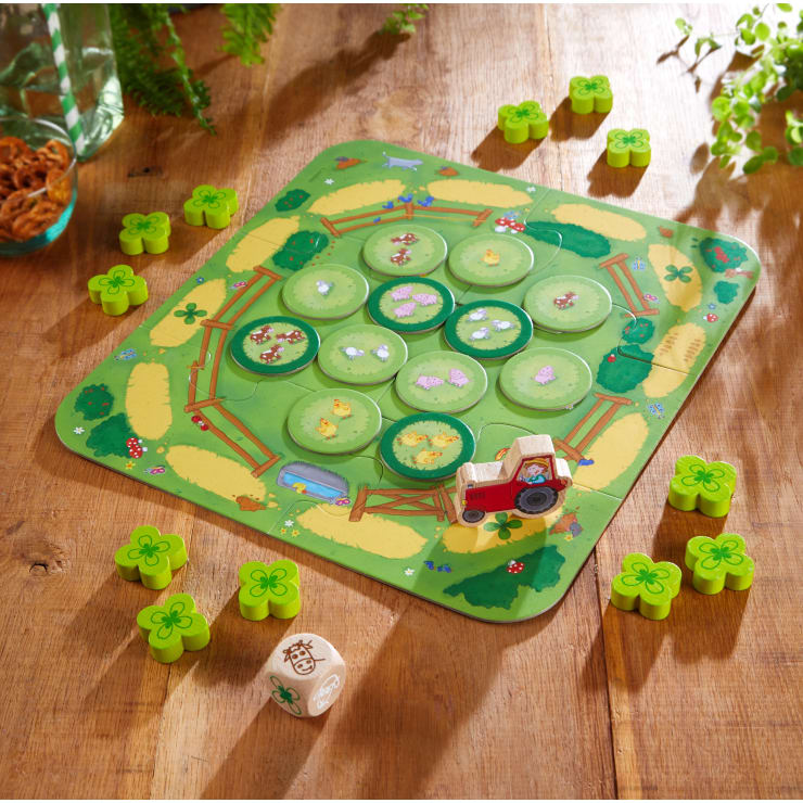Ein farbenfrohes Brettspiel mit einem grünen Spielbrett mit Bauernhofmotiven, runden grünen Spielsteinen, einem roten Traktor, mehreren kleeförmigen Spielsteinen und einem Holzwürfel auf einem Holztisch. Im Hintergrund sind einige Pflanzen und Getreide zu sehen.