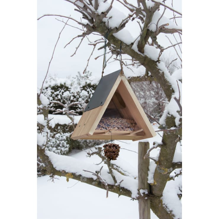 Ein dreieckiges, mit Samen gefülltes Vogelfutterhaus aus Holz hängt an einem schneebedeckten Ast. Ein Tannenzapfen hängt unterhalb des Futters. Schnee bedeckt die umliegenden Äste und den Boden.