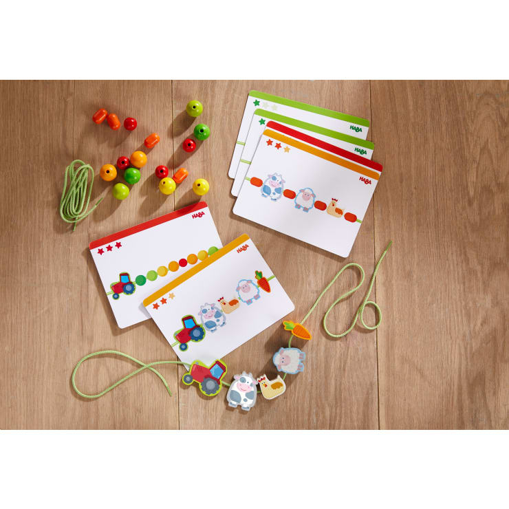 Ein Satz bunter Aktivitätskarten, Holzperlen, Tier- und Fahrzeugformen und grüne Schnürsenkel werden auf einem Holzboden ausgebreitet und sind für die Aktivitäten der Kinder zum Schnüren und Erkennen von Mustern gedacht.