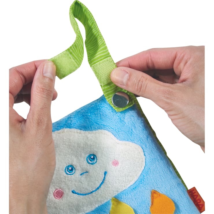Zwei Hände befestigen ein grünes Stoffband mit Klettverschluss an einem weichen, blauen, strukturierten Gegenstand, auf dem eine weiße, lächelnde Wolke mit leuchtend blauen Augen und darunter gestickten orangen Formen zu sehen ist.