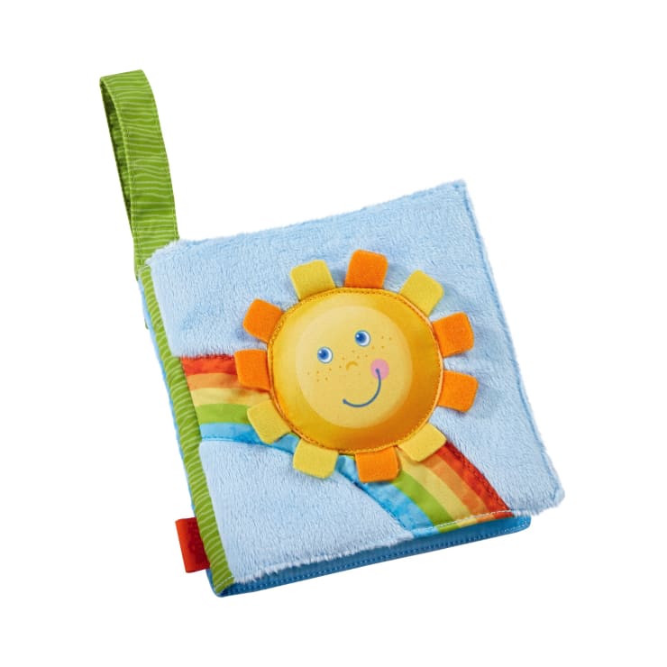 Ein weiches Babybuch aus Stoff mit einem blauen Einband, auf dem eine lächelnde gelbe Sonne und ein Regenbogenmotiv abgebildet sind. Das Buch hat eine grün-gestreifte Stoffschlaufe zur Befestigung.