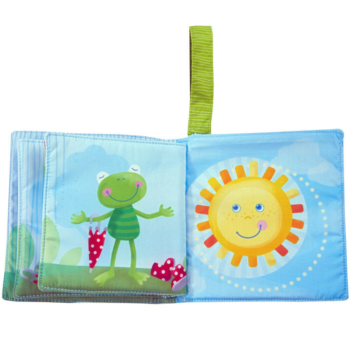 Ein weiches Babybuch aus Stoff ist aufgeschlagen und zeigt einen lächelnden grünen Frosch im Gras mit Pilzen und eine fröhliche Sonne mit einem Gesicht vor einem blauen Himmel. Am oberen Rand ist eine grüne Stoffschleife angebracht.