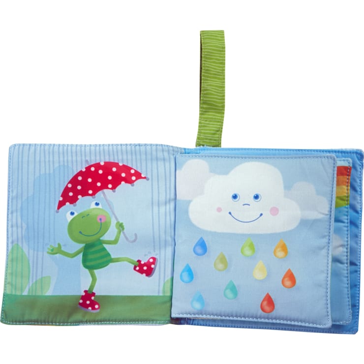 Ein Kinderbuch aus Stoff ist aufgeschlagen und zeigt auf der linken Seite einen grünen Cartoon-Frosch mit einem roten Regenschirm und auf der rechten Seite eine lächelnde weiße Wolke mit bunten Regentropfen. Oben ist eine grün gestreifte Stoffschleife angebracht.