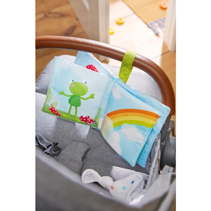 Ein Babybuch aus weichem Stoff mit Illustrationen eines grünen Froschs und eines Regenbogens ist an dem braunen Griff eines grauen Kinderwagens befestigt. Auf dem Boden im Hintergrund sind einige Spielzeuge zu sehen.