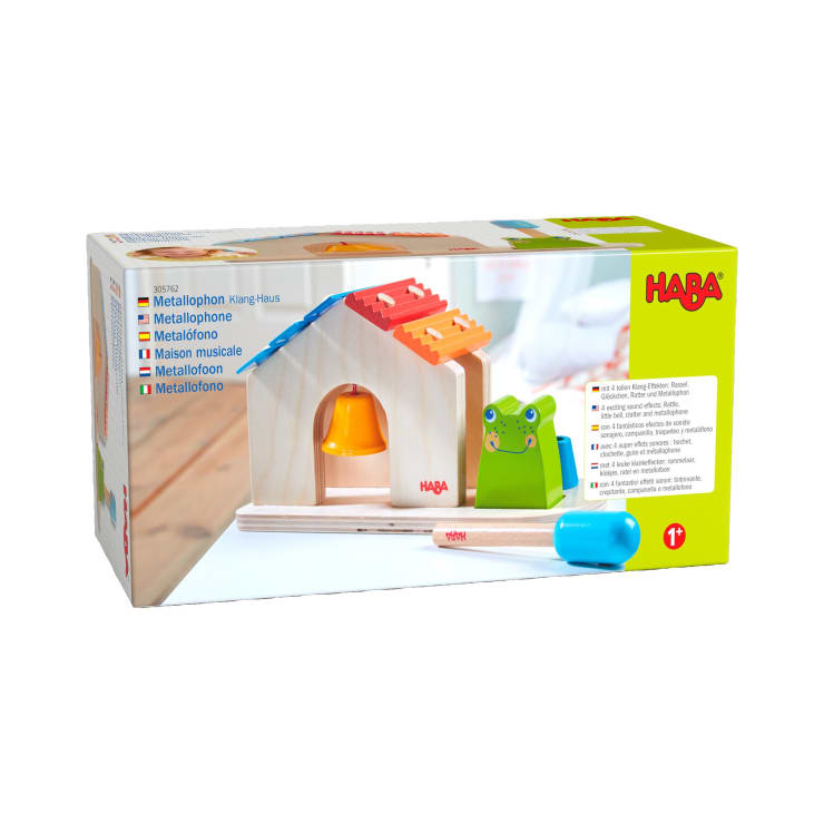 Ein HABA-Spielzeugset in einer Schachtel mit einem Holzhaus mit einer gelben Glocke, Xylophonstäben aus Metall auf dem Dach, einer grünen Froschfigur und zwei Schlägeln. Die Verpackung zeigt Branding und Produktdetails in mehreren Sprachen.