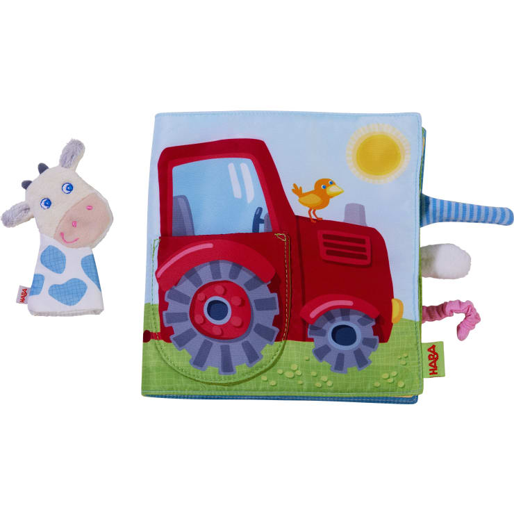 Ein Kinderbuch aus weichem Stoff, das einen roten Traktor mit einem gelben Vogel neben einer blau-weißen Plüschkuh-Fingerpuppe zeigt. Das Buch hat Stofflaschen und eine Sonnenillustration im Hintergrund.