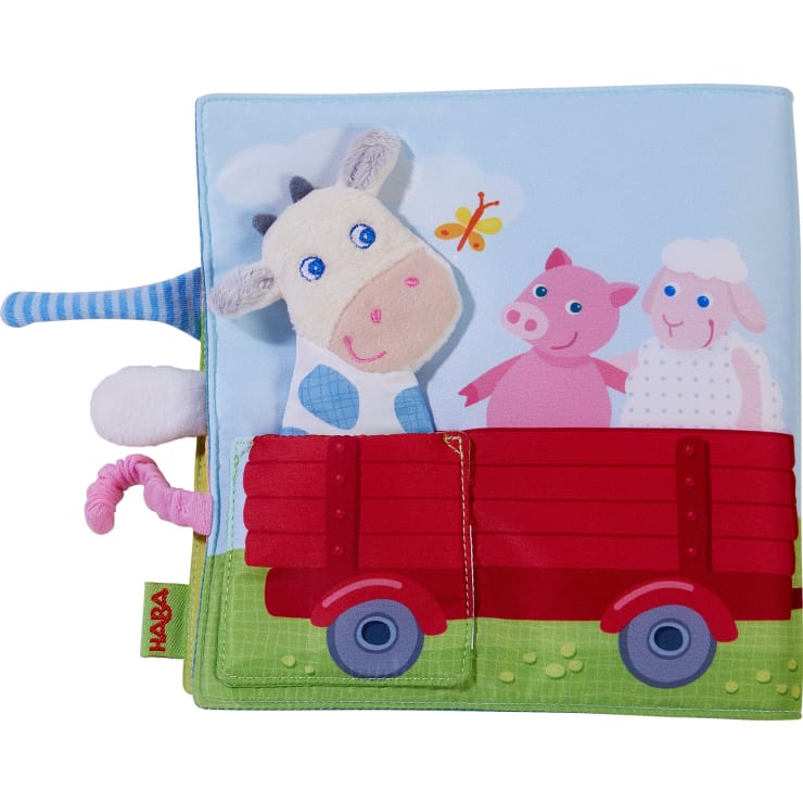 Ein Kinderbuch aus Stoff mit einer Kuhpuppe, einem Schwein und einem Schaf in einem roten Wagen auf einer grünen Wiese, mit einem blauen Himmel und einem gelben Schmetterling im Hintergrund. Das Buch hat verschiedene strukturierte Elemente.