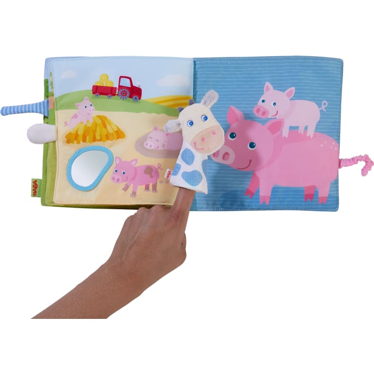 Eine Hand hält eine Kuh-Fingerpuppe über einem aufgeschlagenen Stoffbuch. Die linke Seite zeigt eine Bauernhofszene mit einem Traktor, Heu und einem Spiegel. Auf der rechten Seite sind drei rosa Schweine auf blauem Hintergrund abgebildet.