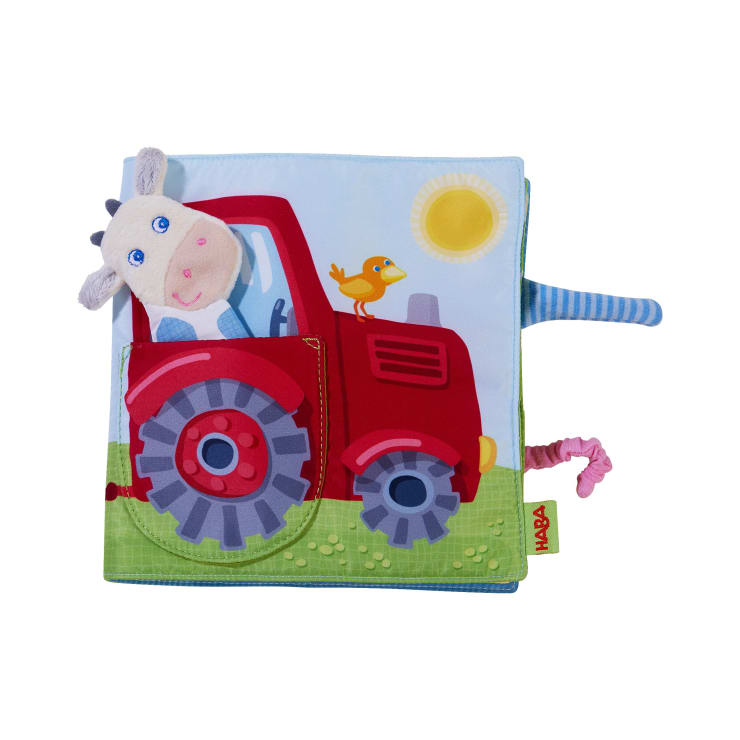 Ein weiches Stoff-Kinderbuch mit einem roten Traktor, einer Plüschkuh auf dem Fahrersitz und einem gelben Vogel auf der Motorhaube. Der Hintergrund zeigt blauen Himmel, Sonne und grünes Gras. Verschiedene Stofflaschen ragen aus dem Buch heraus.