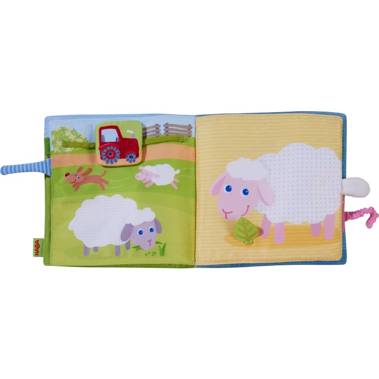 Ein buntes Kinderbuch aus Stoff zeigt Cartoon-Schafe, einen roten Traktor, einen Hund, der ein Lamm jagt, und ein großes Schaf mit einem rosa Gesicht, das ein Blatt frisst. Der Schwanz des Schafes ist ein weißer, flauschiger Stoffknubbel.