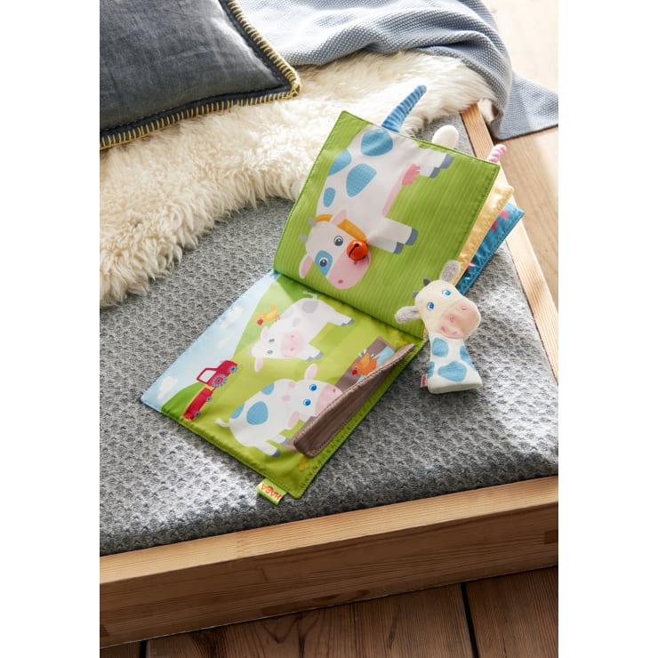 Ein Kinderbuch aus weichem Stoff mit bunten Illustrationen von Kühen ist auf einer grauen, strukturierten Oberfläche neben einem Plüschkuh-Spielzeug aufgeschlagen. Die Szene ist auf einem Holzbett mit einer Fleecedecke und einem grauen Kissen zu sehen.