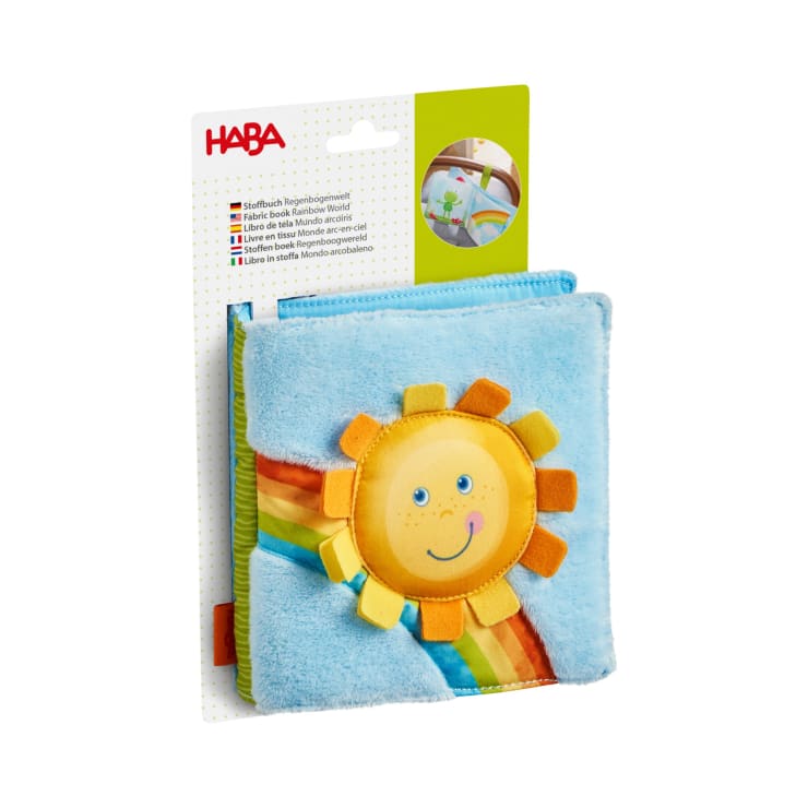 Ein Babybuch aus weichem Stoff mit dem Titel Rainbow World von HABA, mit einem plüschigen blauen Einband mit einer lächelnden Sonne und einem Regenbogenmotiv, in seiner Verpackung mit Produktinformationen in mehreren Sprachen.