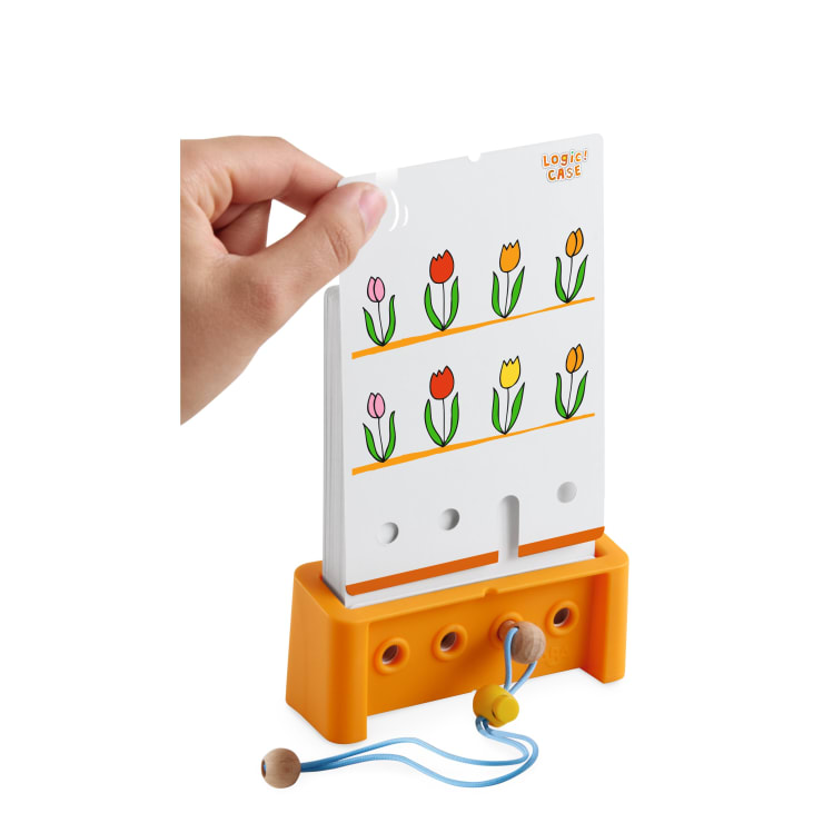Eine Hand hebt eine weiße Karte mit Tulpenzeichnungen aus einem orangefarbenen Ständer mit der Aufschrift LOGIC! KASTEN. Der Ständer hat Löcher, eine Schnur und eine Holzperle an der Vorderseite.