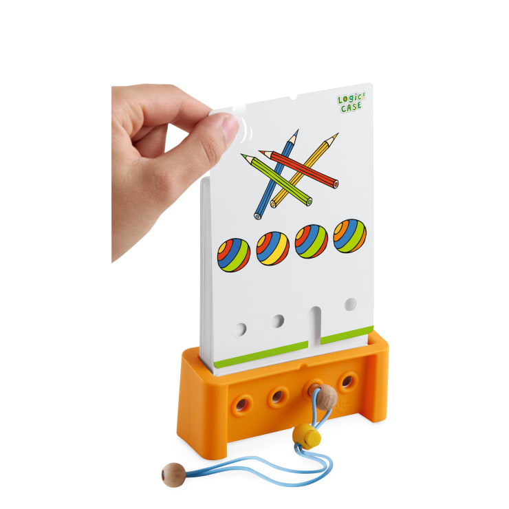 Eine Hand hebt eine Karte mit Abbildungen von vier Buntstiften und drei gestreiften Kugeln von einem orangefarbenen Plastikständer. Am Ständer ist eine blaue Kordel mit einer Holzperle befestigt.