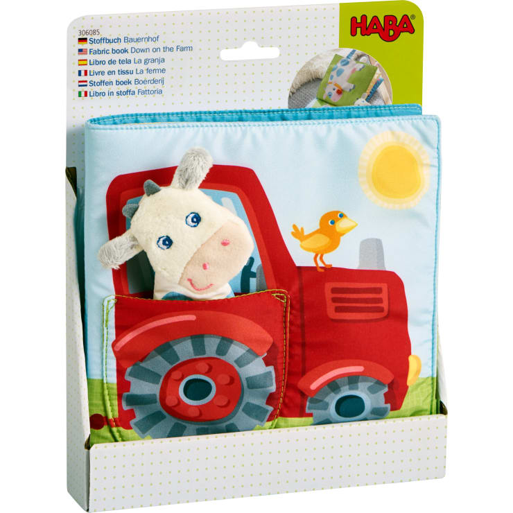 Ein Stoffbuch mit dem Titel Auf dem Bauernhof von HABA wird in seiner Verpackung gezeigt. Das Cover zeigt eine ausgestopfte Kuh in einem roten Traktor mit einem gelben Vogel auf der Motorhaube und einer aufgedruckten Sonne am Himmel.