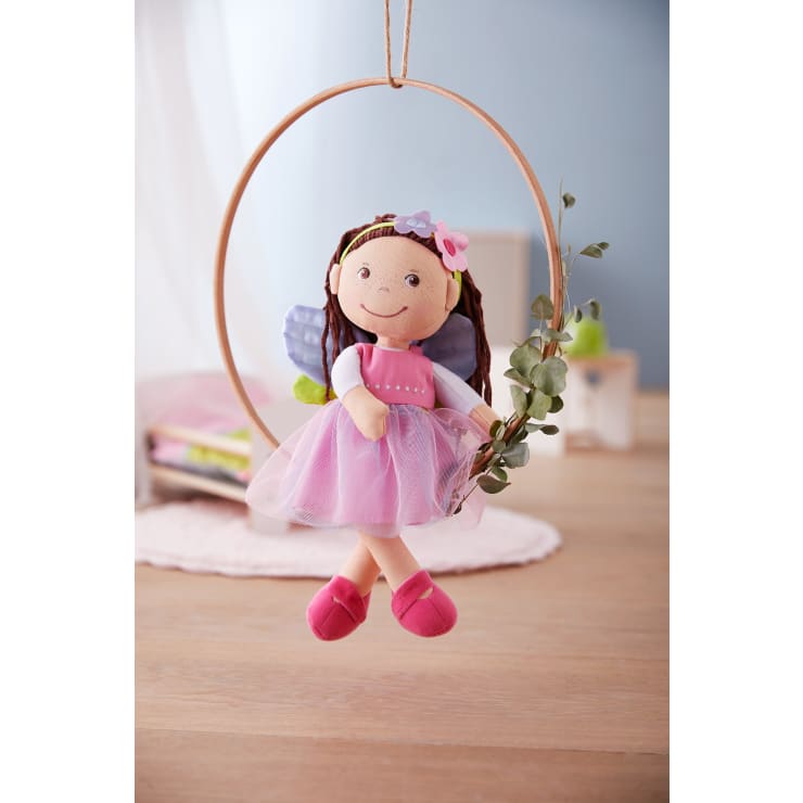 Eine Feenpuppe aus Stoff mit braunem Garnhaar, rosa Kleid, Flügeln und Blumenstirnband sitzt in einem mit Grünzeug geschmückten Holzreifen. Der Hintergrund ist ein sanft beleuchteter Raum mit hellen Möbeln und Dekor.