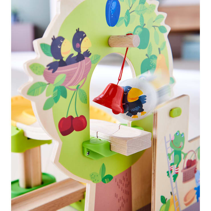 Ein Holzspielzeug mit bunten Illustrationen von Vögeln, Kirschen und einem Frosch. Ein kleiner Ball mit einem Vogelmotiv schwingt an einer Schnur und schlägt gegen eine rote Glocke, die am Rahmen des Spielzeugs befestigt ist.