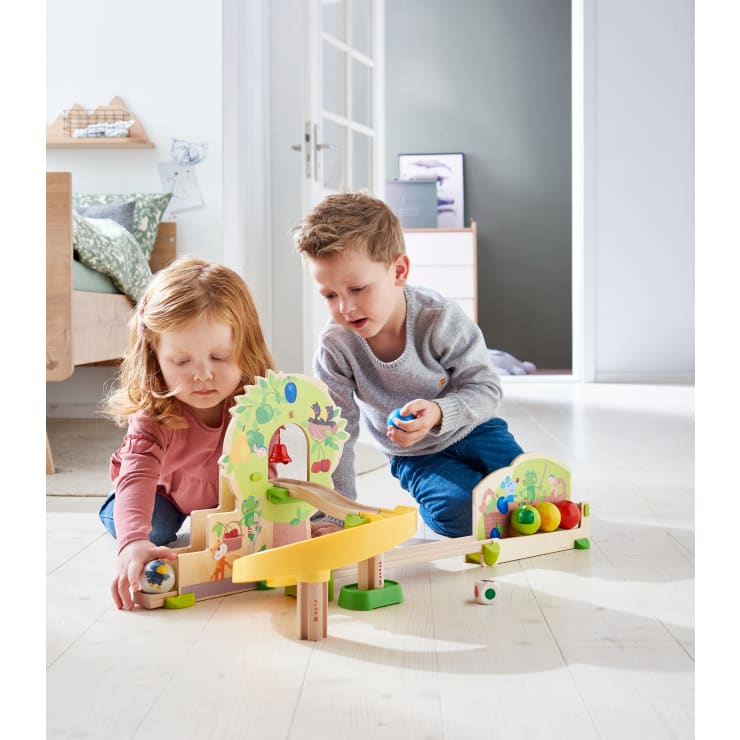 Zwei kleine Kinder, ein Mädchen und ein Junge, sitzen auf einem hellen Holzboden und spielen mit einem Holzspielzeugset mit Rampen, Blöcken und bunten Bällen in einem hellen, modernen Raum.